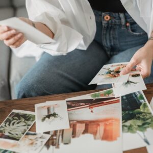 Crop unrecognizable tattooed female sitting on sofa at desk with assorted photos of flowers and town in house room